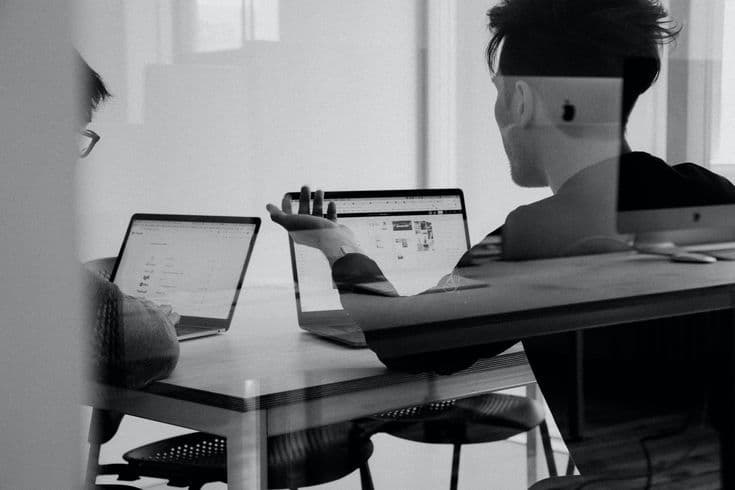 Two colleagues discussing work at laptops across a table, seen through glass